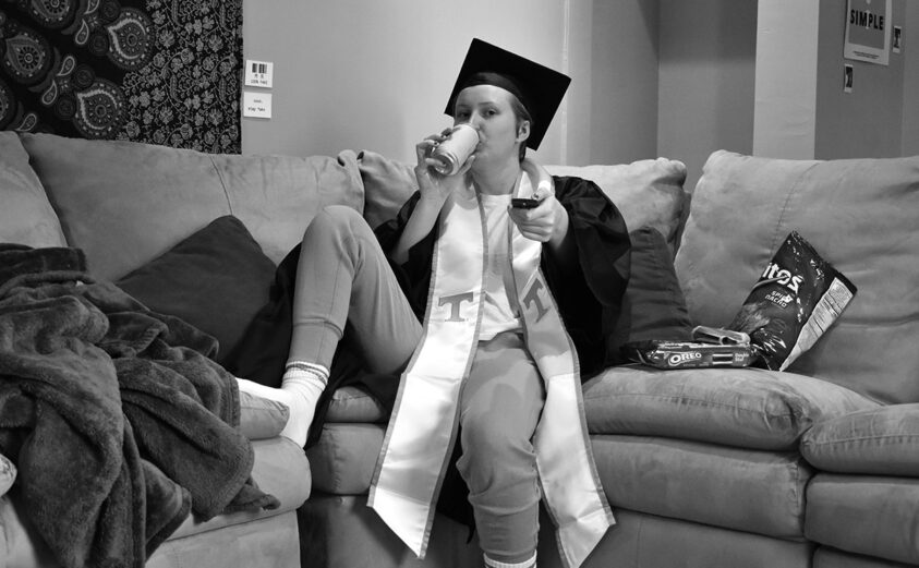 Image of a student on their couch, with their graduation attire on, enjoying a soda and holding their TV remote and snacks.