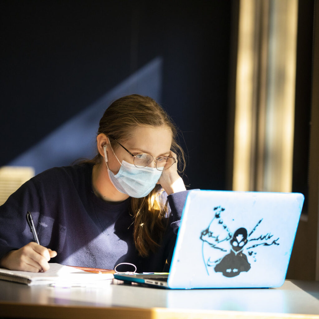 A female student with a mask and a laptop is taking notes on a notebook 